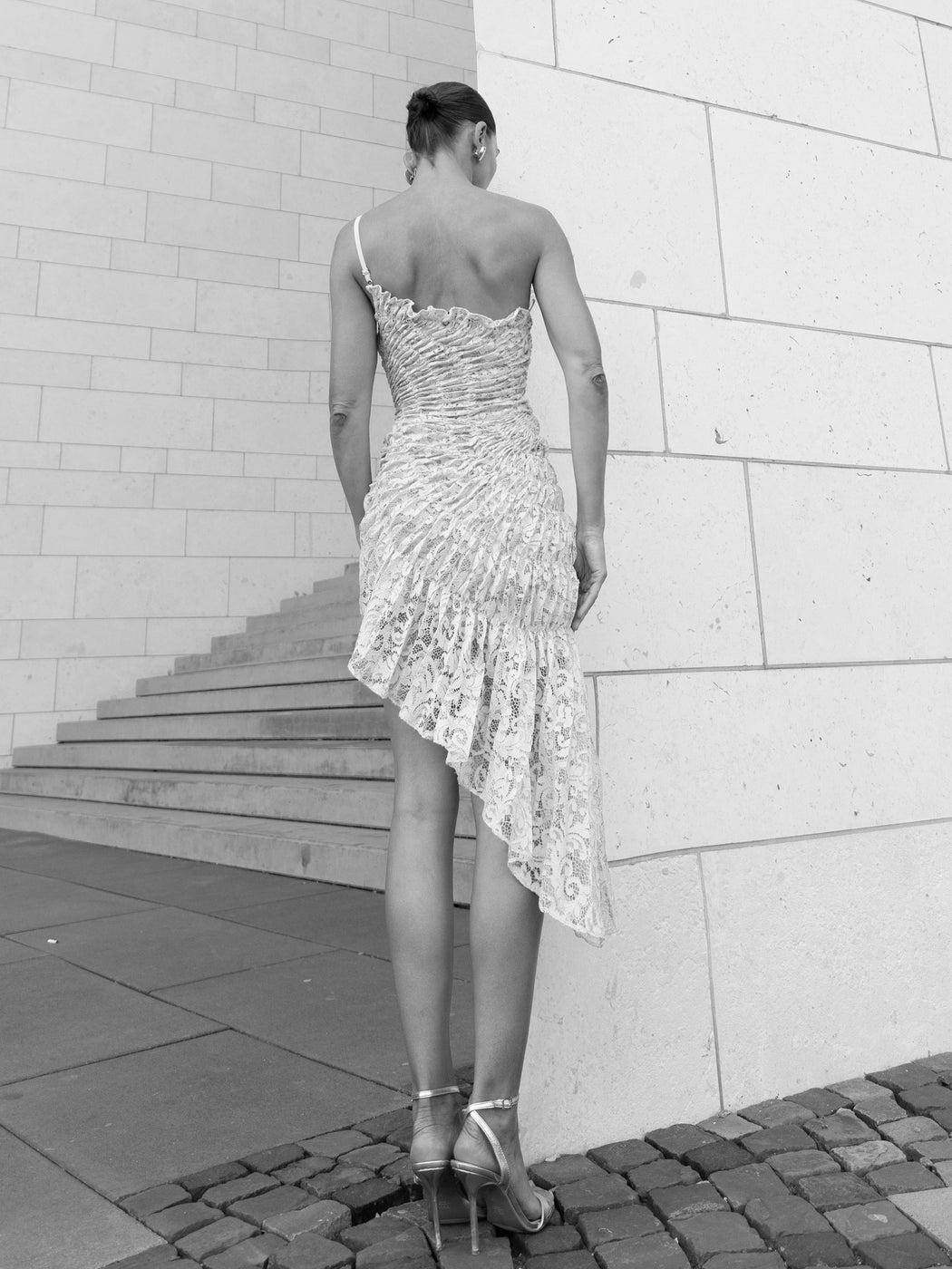 Black and white photo of model in ivory coloured sleeveless dress pleated in a sunray pattern with french baroque style lace facing a wall.