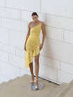 Model wearing a yellow one-shoulder dress featuring a fossil inspired design, pleated in a sunray pattern on French baroque style lace, standing against a white tiled wall.