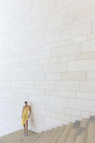 Model in a lemon-coloured sleeveless dress pleated in a sunray pattern with French baroque style lace standing on a staircase against a white wall.