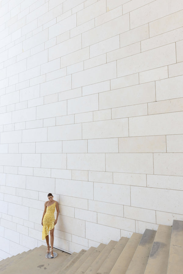 Model in a lemon-coloured sleeveless dress pleated in a sunray pattern with French baroque style lace standing on a staircase against a white wall.