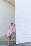 Model wearing a pink one-shoulder dress featuring a fossil inspired design, pleated in a sunray pattern on French baroque style lace, standing against a white wall.