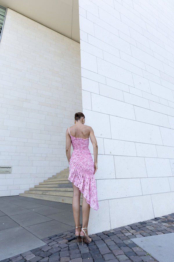 Model wearing a pink one-shoulder dress featuring a fossil inspired design, pleated in a sunray pattern on French baroque style lace, standing against a white brick wall.