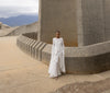 Model in a white floor-length dress pleated in flowing waterfall design standing in front of a large concrete structure.