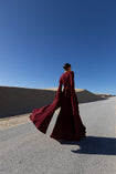 Model in a merlot coloured floor  length dress pleated in flowing waterfall design with wide sleeves and high neckline back turned while standing on a road with sand dunes and blue sky in the background