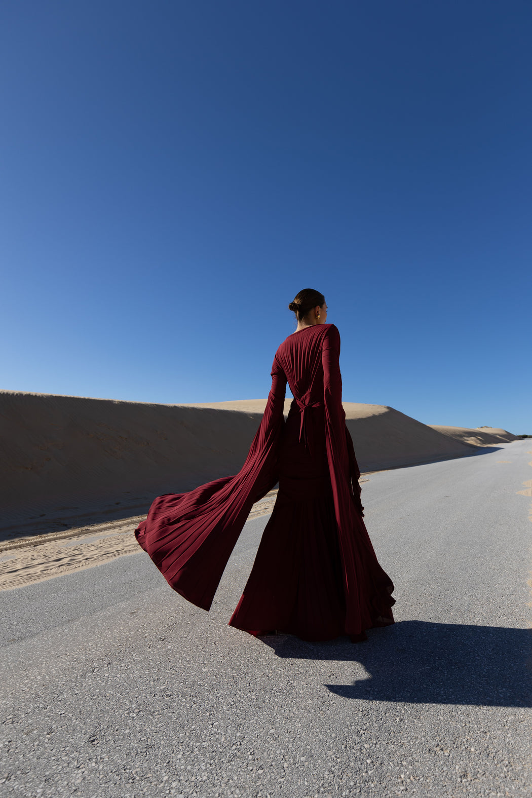 Model in a merlot coloured floor  length dress pleated in flowing waterfall design with wide sleeves and high neckline back turned while standing on a road with sand dunes and blue sky in the background