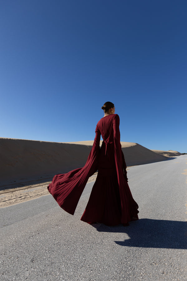 Model in a merlot coloured floor  length dress pleated in flowing waterfall design with wide sleeves and high neckline back turned while standing on a road with sand dunes and blue sky in the background