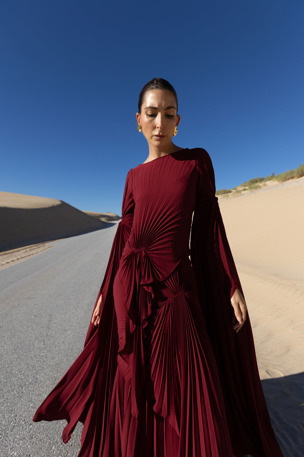 Model in a merlot coloured floor  length dress pleated in flowing waterfall design with wide sleeves and high neckline standing on a road with sand dunes and blue sky in the background