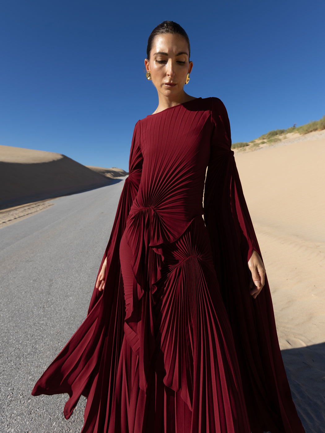 Model in a merlot coloured floor  length dress pleated in flowing waterfall design with wide sleeves and high neckline standing on a road with sand dunes and blue sky in the background.