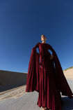 Model in a merlot coloured floor  length dress pleated in flowing waterfall design with wide sleeves and high neckline, looking down while standing on a road with sand dunes and blue sky in the background