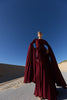 Model in a merlot coloured floor  length dress pleated in flowing waterfall design with wide sleeves and high neckline, looking down while standing on a road with sand dunes and blue sky in the background