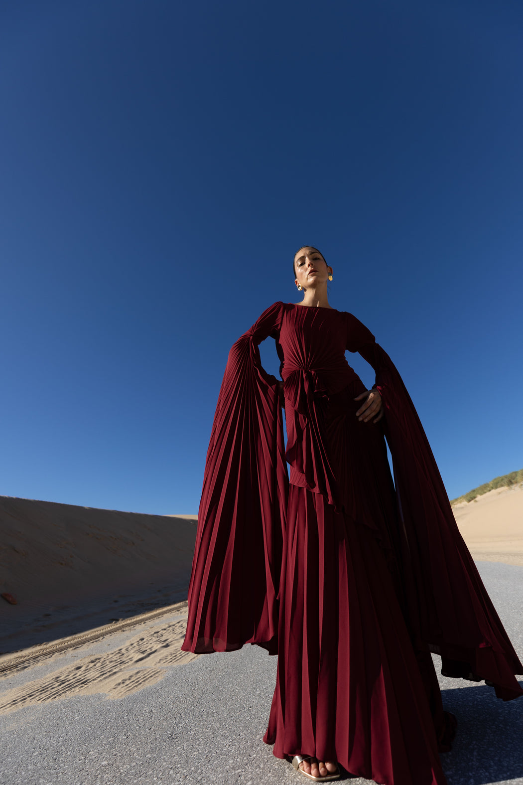 Model in a merlot coloured floor  length dress pleated in flowing waterfall design with wide sleeves and high neckline, looking down while standing on a road with sand dunes and blue sky in the background
