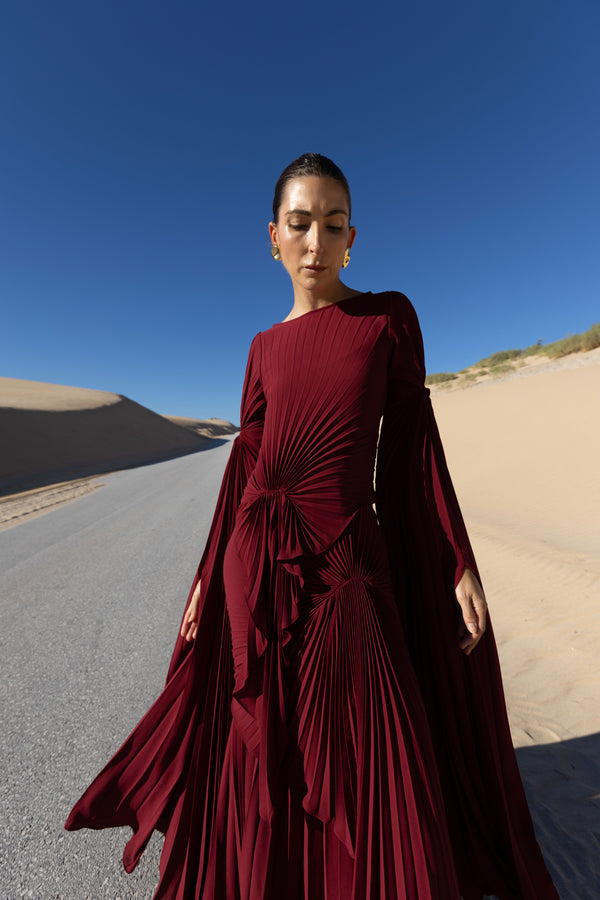 Model in a merlot coloured floor  length dress pleated in flowing waterfall design with wide sleeves and high neckline standing on a road with sand dunes and blue sky in the background