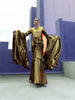 Model in a gold liquid metallic floor-length dress pleated in a flowing waterfall design with long sleeves standing in front of a futuristic cityscape