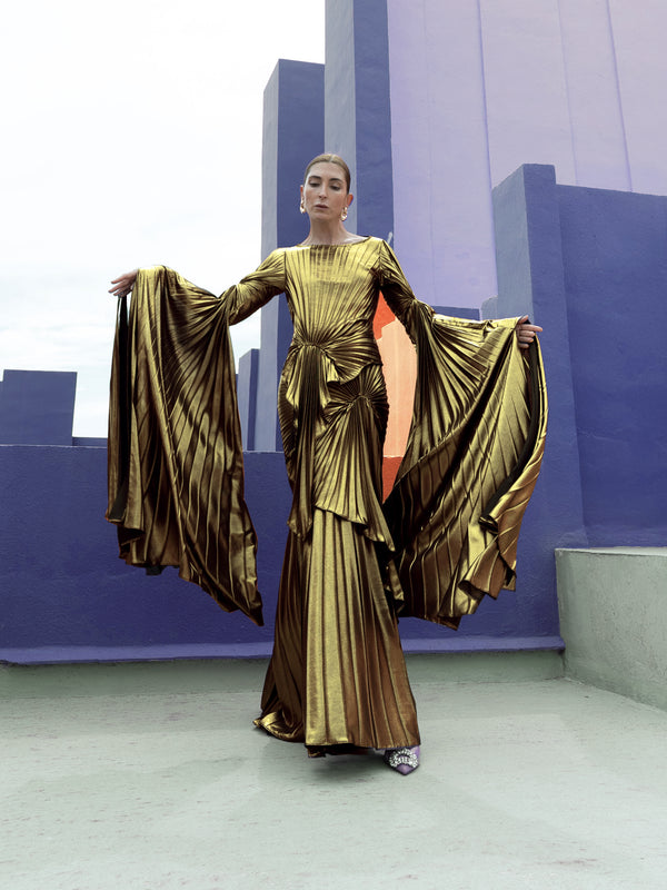Model in a gold liquid metallic floor-length dress pleated in a flowing waterfall design with long sleeves standing in front of a futuristic cityscape