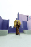Model in a Gold liquid metallic floor-length dress pleated in a flowing waterfall design with long sleeves standing in a purple futuristic cityscape