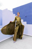 Model in a Gold liquid metallic floor-length dress pleated in a flowing waterfall design with long sleeves standing against a blue and white abstract wall.