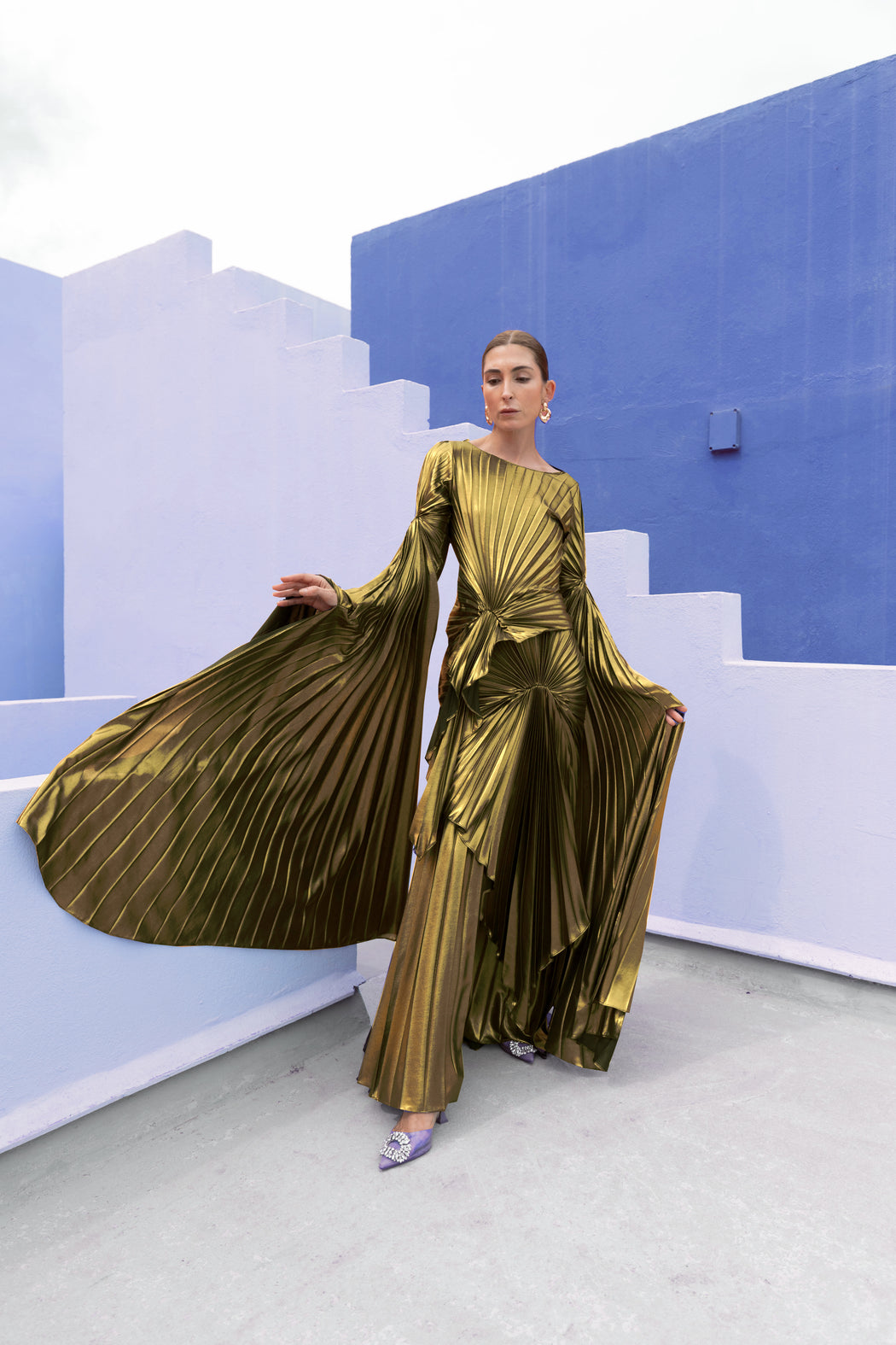 Model in a Gold liquid metallic floor-length dress pleated in a flowing waterfall design with long sleeves standing against a blue and white abstract wall.