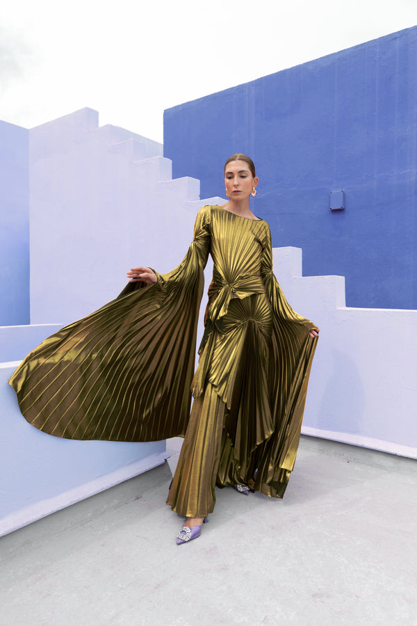 Model in a Gold liquid metallic floor-length dress pleated in a flowing waterfall design with long sleeves standing against a blue and white abstract wall.