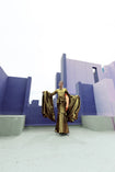 Model in a gold liquid metallic floor-length dress pleated in a flowing waterfall design with long sleeves standing in front of a geometric architectural structure.