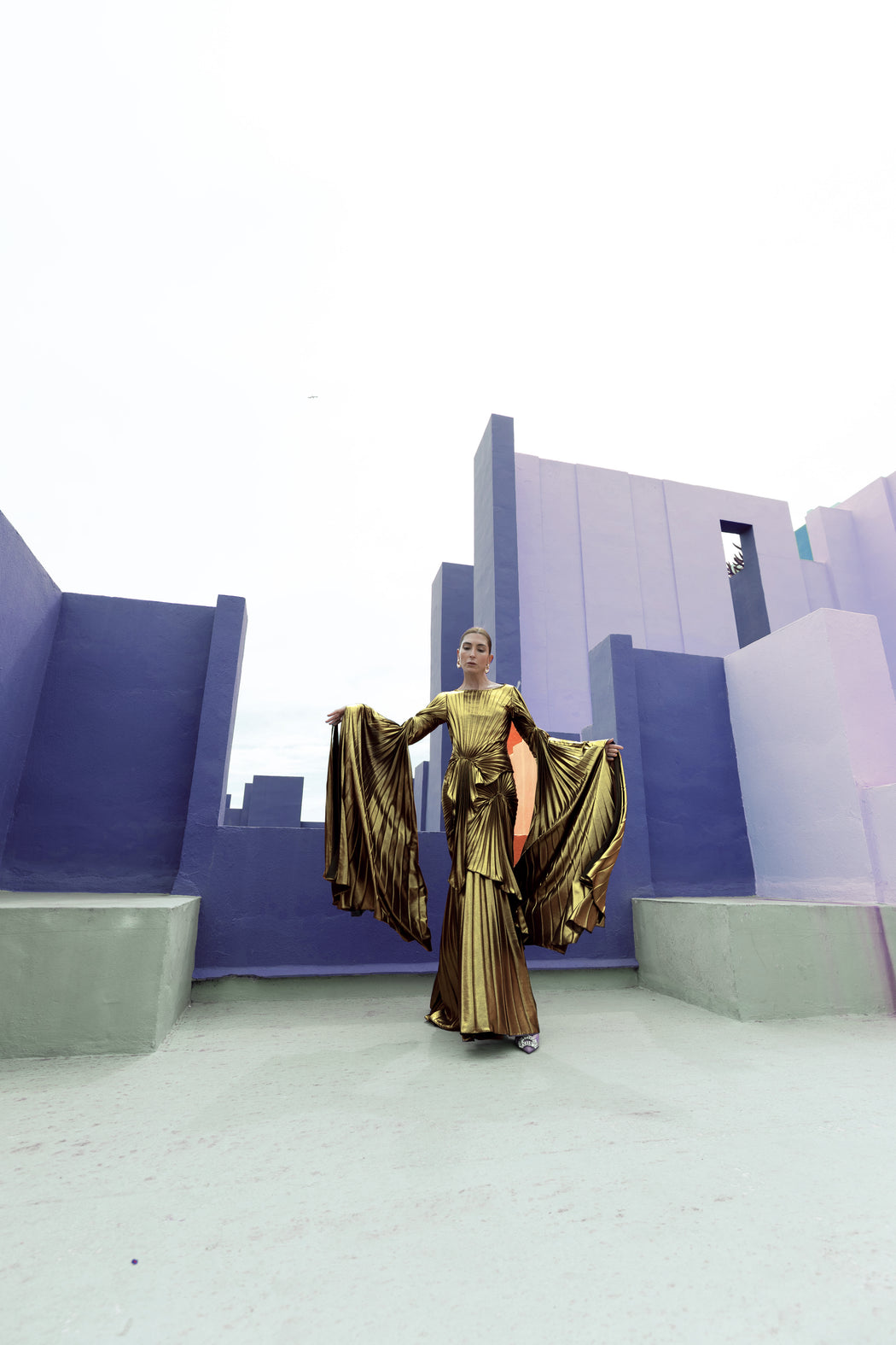 Model in a gold liquid metallic floor-length dress pleated in a flowing waterfall design with long sleeves standing in front of a geometric architectural structure.
