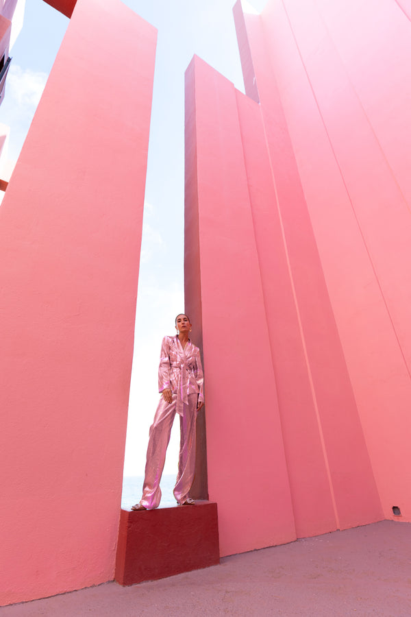 Model wearing a pink wrap-style sequin blazer with sunray stripes and matching wide-leg trousers standing between two tall pink structures with a clear blue sky.
