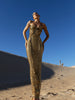Model in a gold pleated ankle-length sequin dress with unique tuck detailing down the front with a separate bamboo jersey lace posing on a sand dune under a clear blue sky.