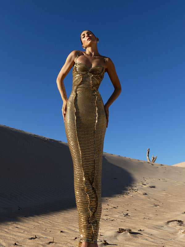 Model in a gold pleated ankle-length sequin dress with unique tuck detailing down the front with a separate bamboo jersey lace posing on a sand dune under a clear blue sky.